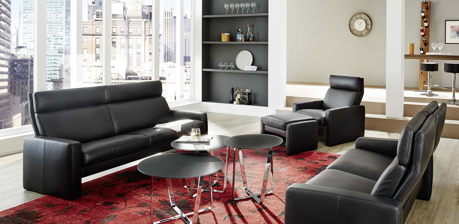 Arosa Couch mit Sessel und Hocker in schwarzem Leder, schwarzen runden Beistelltischen und rotem Teppich.