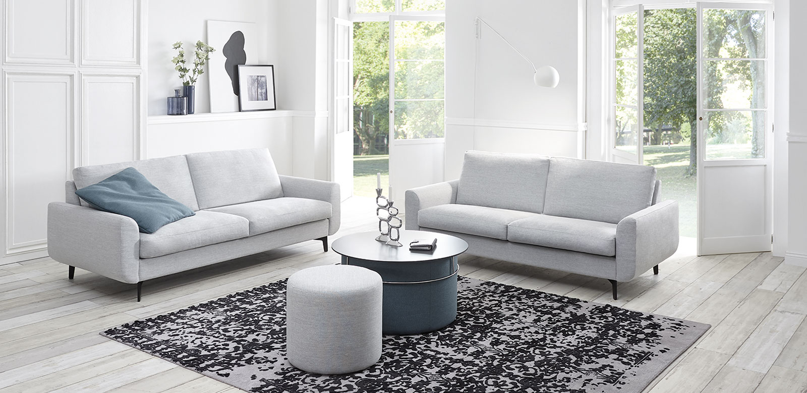 Twee CL830 banken in grijs-witte stof met ronde tafelkrukjes en blauwe kussens in de witte woonkamer van een landhuis